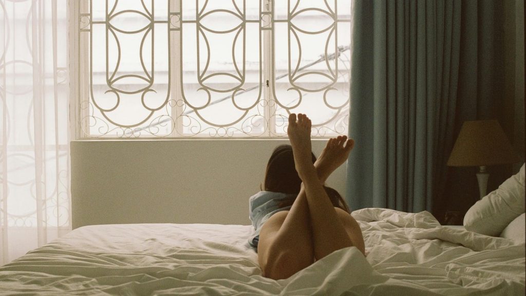 woman in shorts lying in white bed with her feet lifting watching ouside of the window