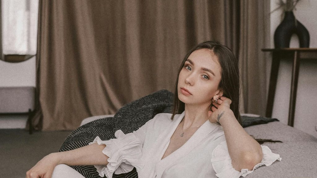 A woman in a white low-necked dress is sitting on the sofa touching her earrings