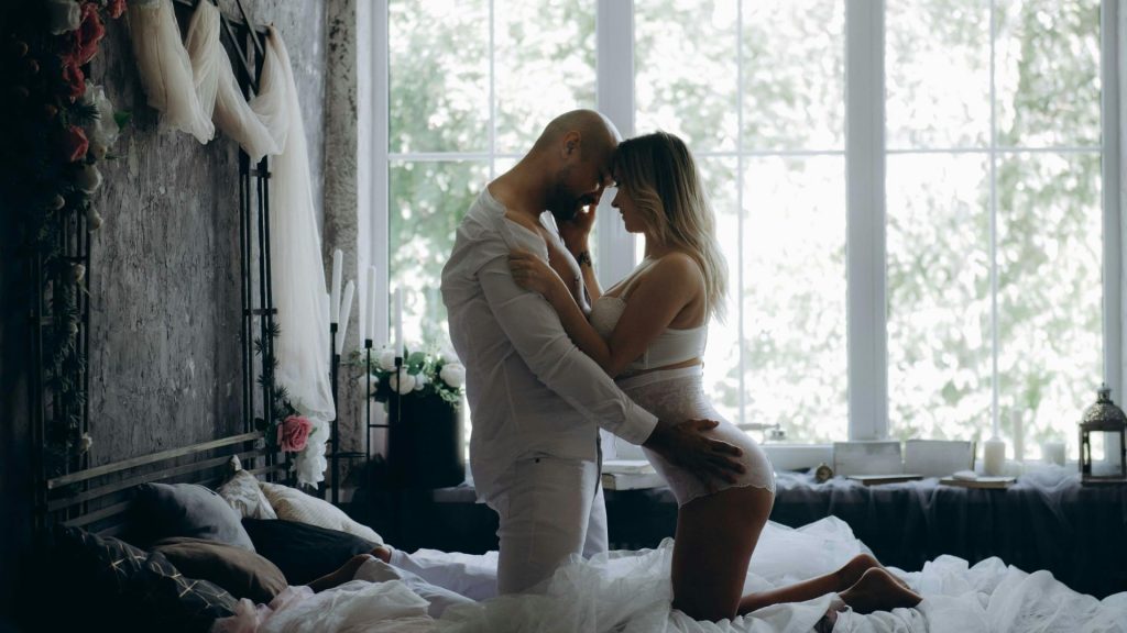 A bald bearded man and a woman in a white lace bra and panties snuggling on the bed